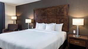 A hotel room with a large bed, two bedside tables with lamps, a chair, and a dark wooden headboard.