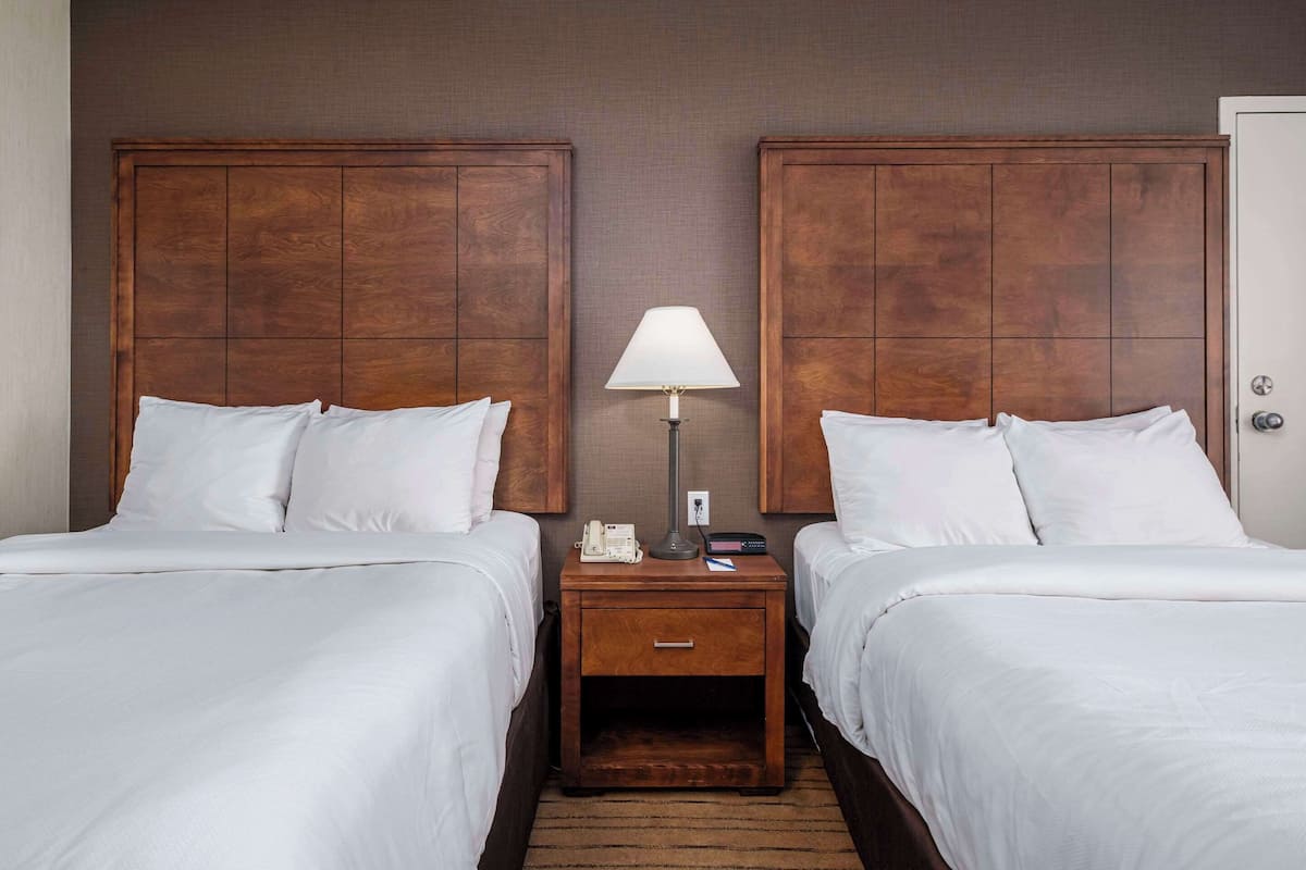 A hotel room with two beds, a nightstand with a lamp, a phone, and a small drawer.