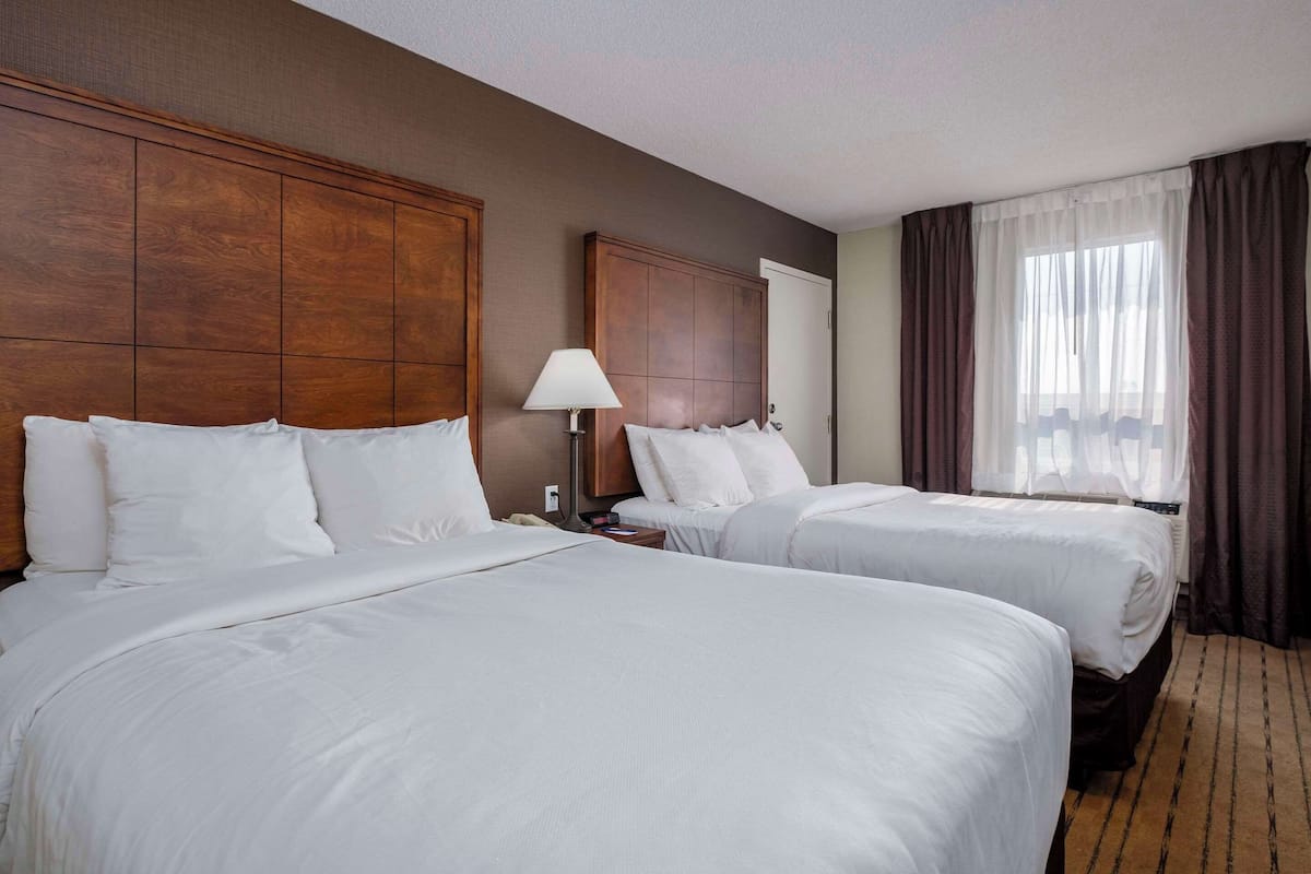 A hotel room with two beds, a wooden headboard, a nightstand with a lamp, and a window with curtains.