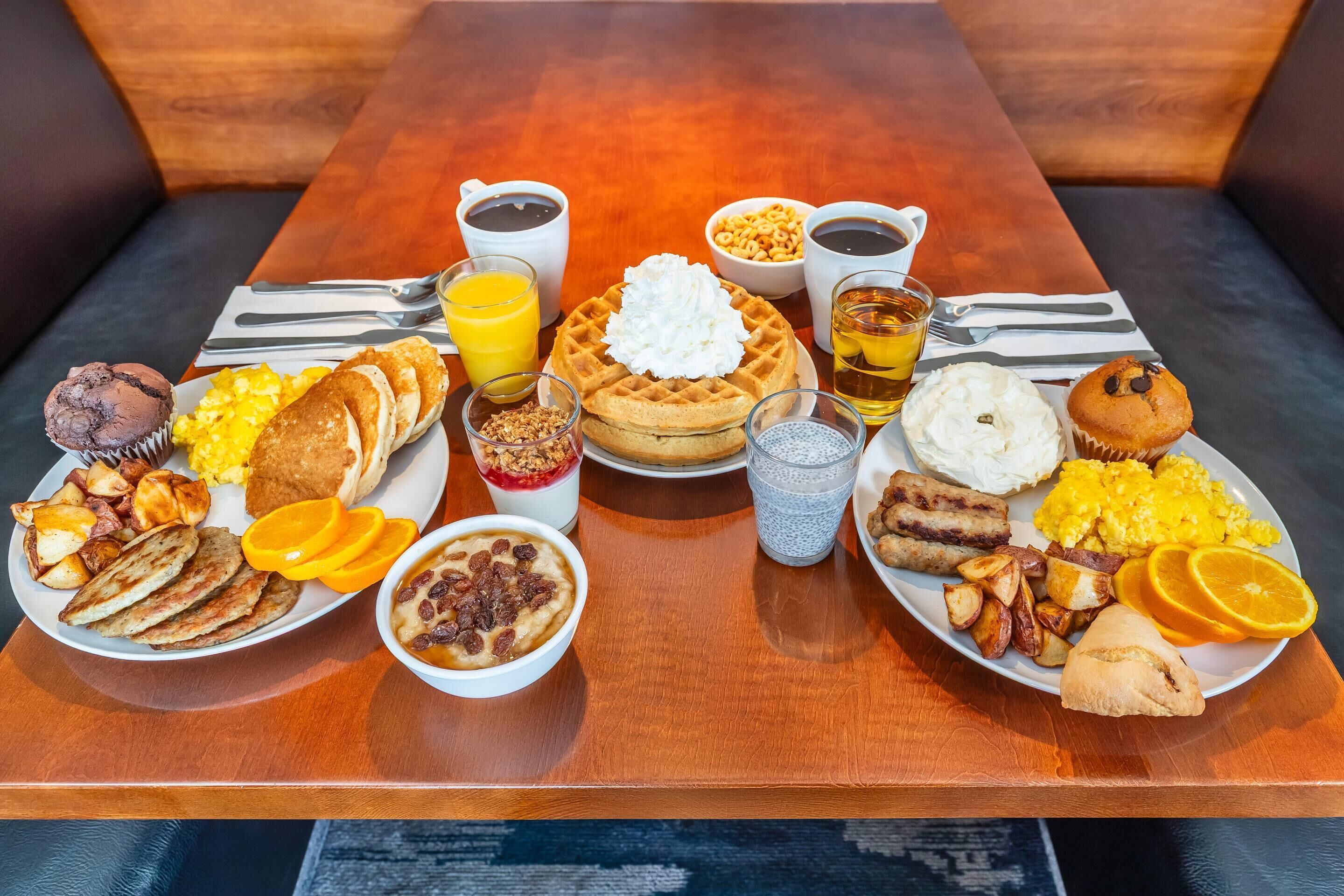 Uma mesa de madeira com um buffet de café da manhã, incluindo panquecas, waffles, ovos e diversos doces.