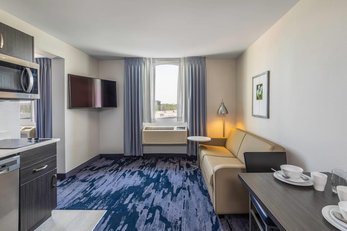 A hotel room with a kitchenette, a flat-screen TV, a sofa, a dining table, and a window with curtains.
