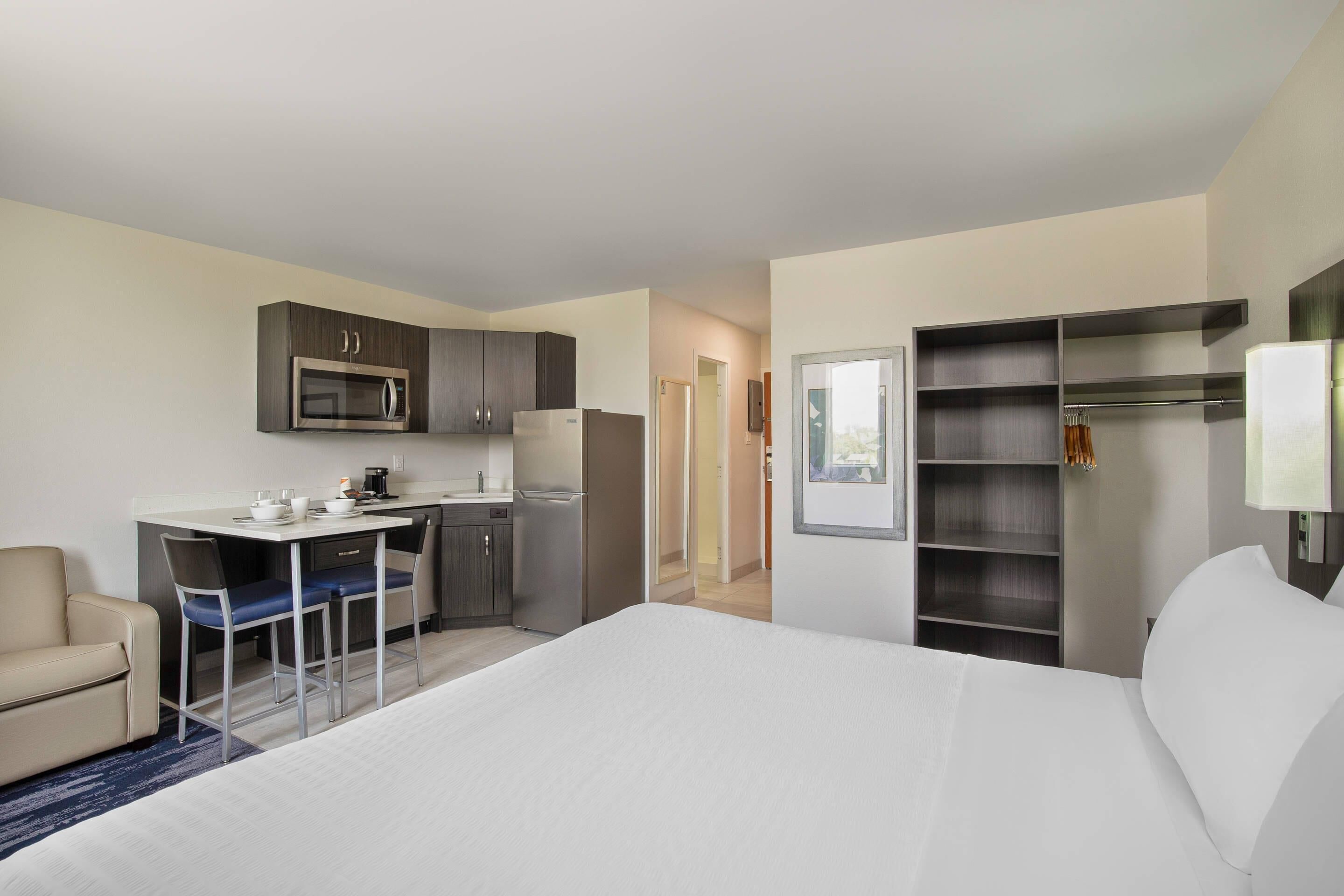 A hotel room with a bed, a desk with chairs, a kitchenette, and a wardrobe.