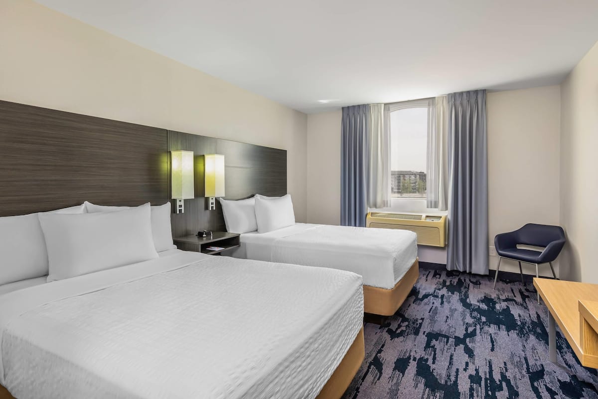 A hotel room with two beds, a desk, a chair, a window with curtains, and a nightstand with lamps.