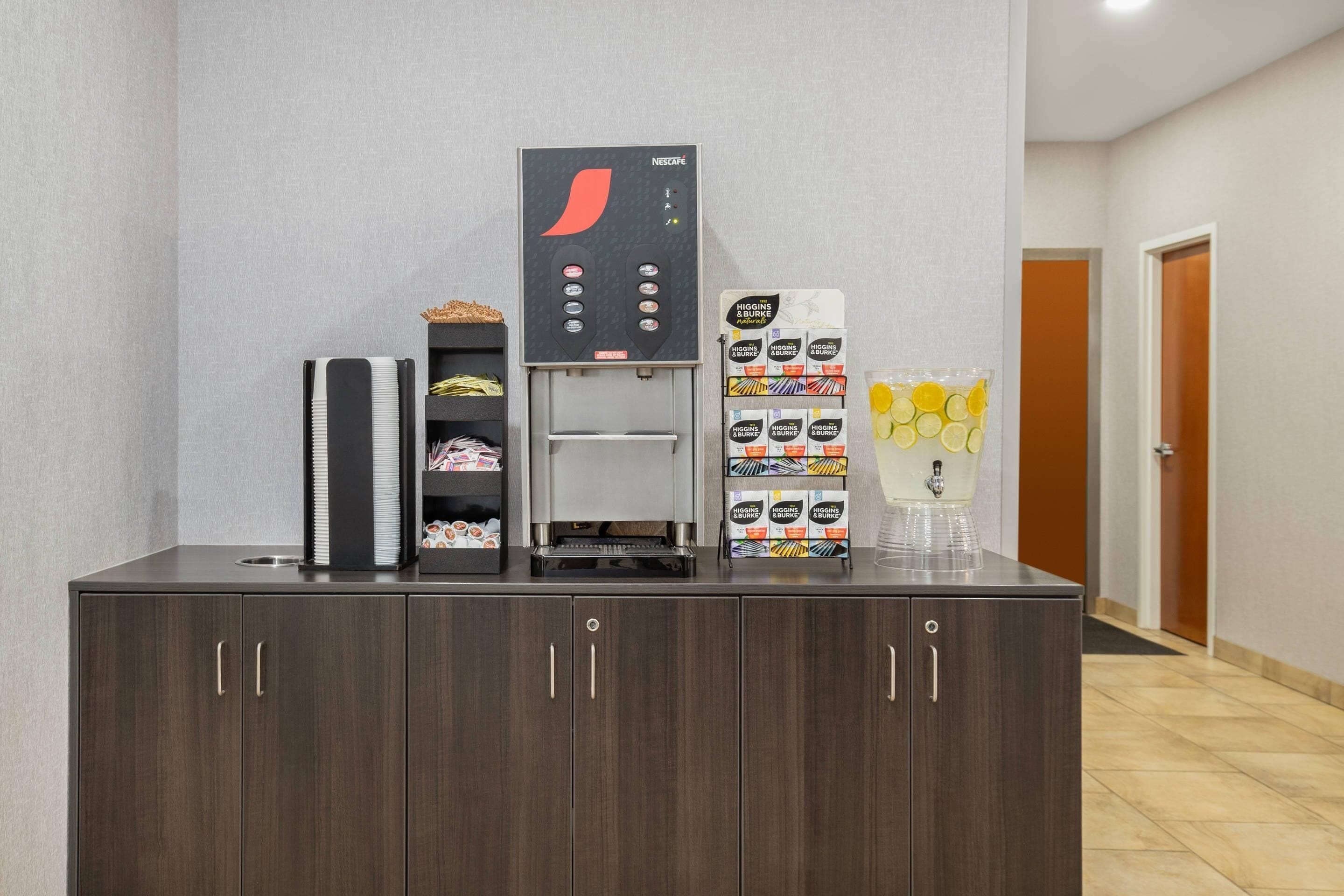 A self-service beverage station with a Nescafe coffee machine, a tea dispenser, and a refrigerated display of beverages.