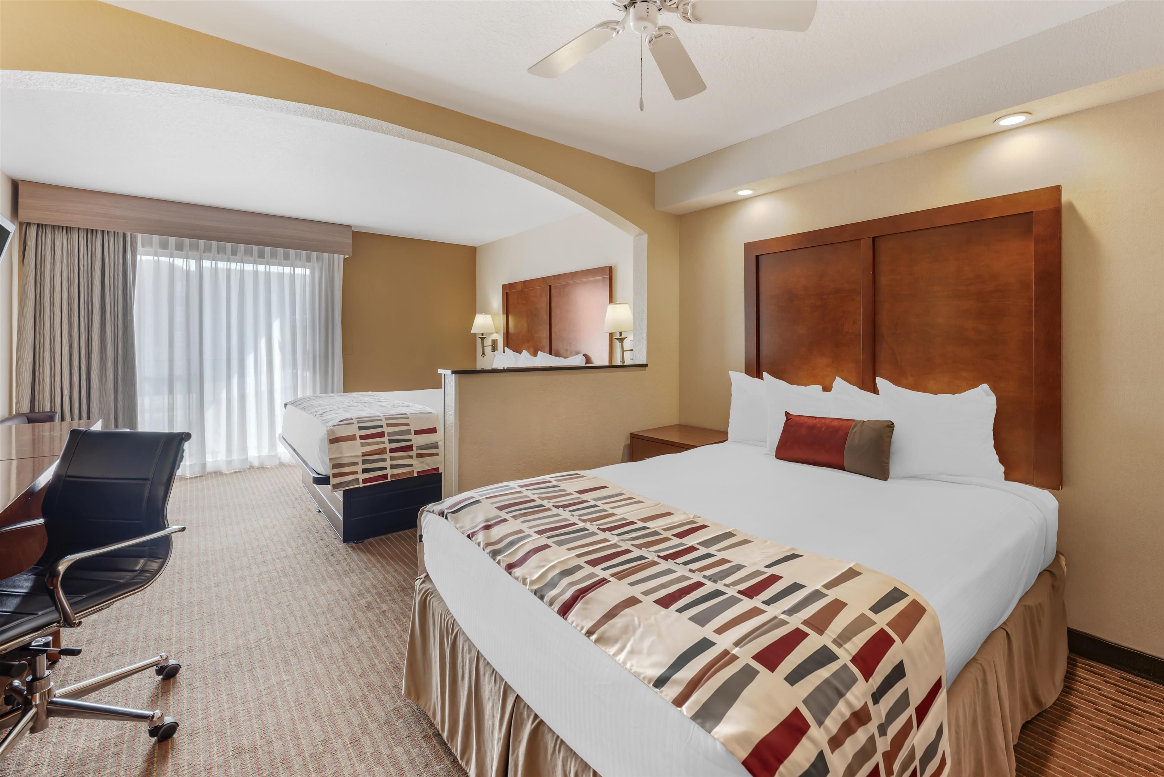 Ein Hotelzimmer mit einem großen Bett, einem Schreibtisch mit Stuhl, einer Schreibtischlampe und einem Deckenventilator.