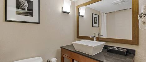 Une salle de bain avec des toilettes blanches, un lavabo avec un meuble en bois, un grand miroir et un tableau encadré au mur.