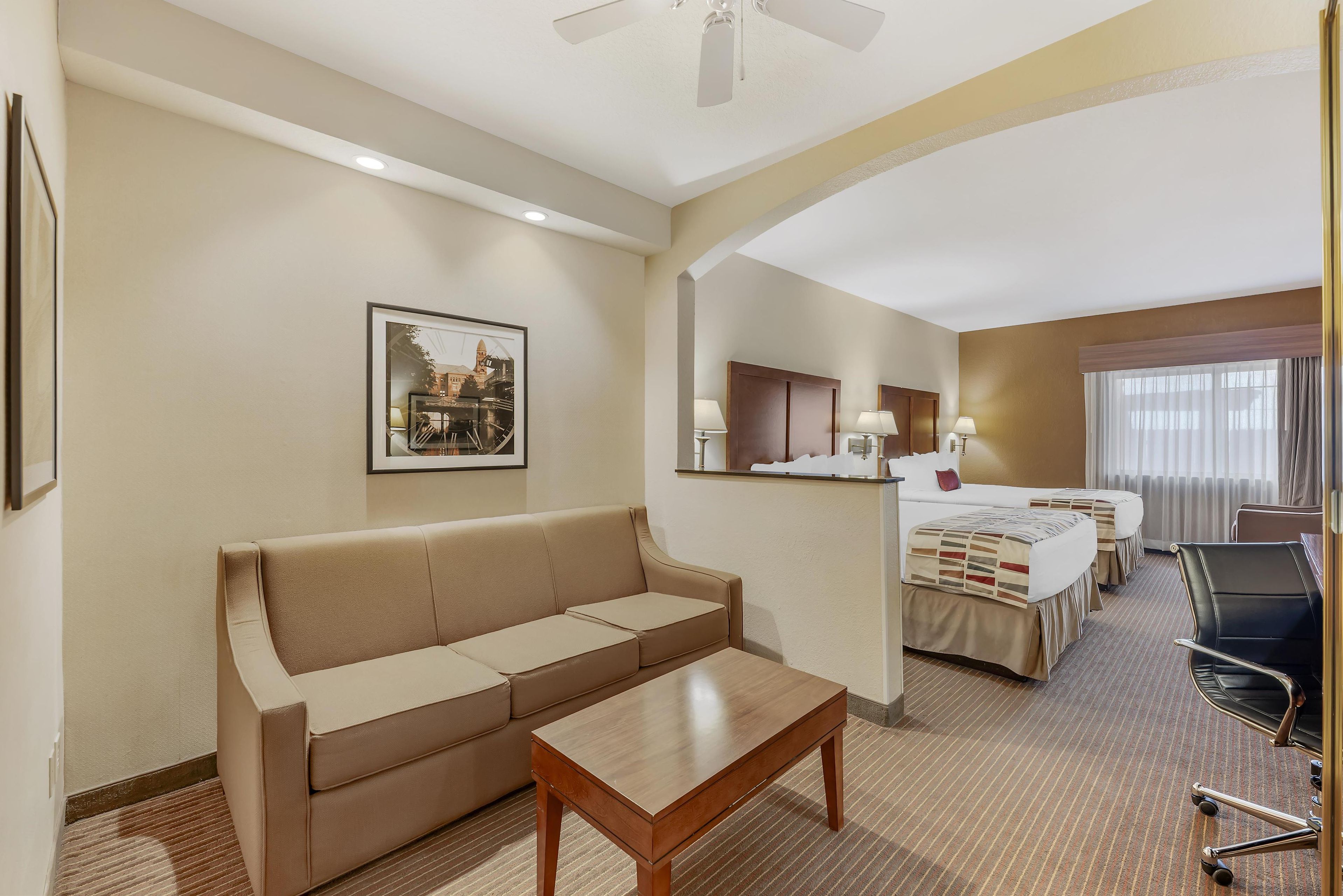 Una habitación de hotel con sofá, una mesita de madera, cama con cabecero, escritorio con silla y ventana con cortinas.