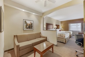 A hotel room with a sofa, a small wooden table, a bed with a headboard, a desk with a chair, and a window with curtains.