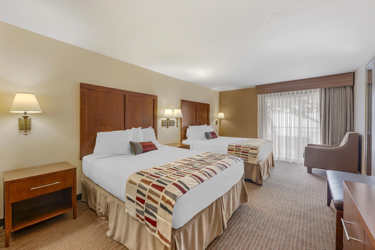 A hotel room with two beds, a desk, a chair, and a large window with curtains.