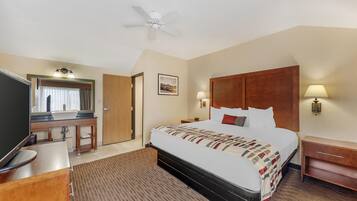 Habitación de hotel con cama, escritorio con espejo, televisión, ventilador de techo y mesita de noche con lámpara.