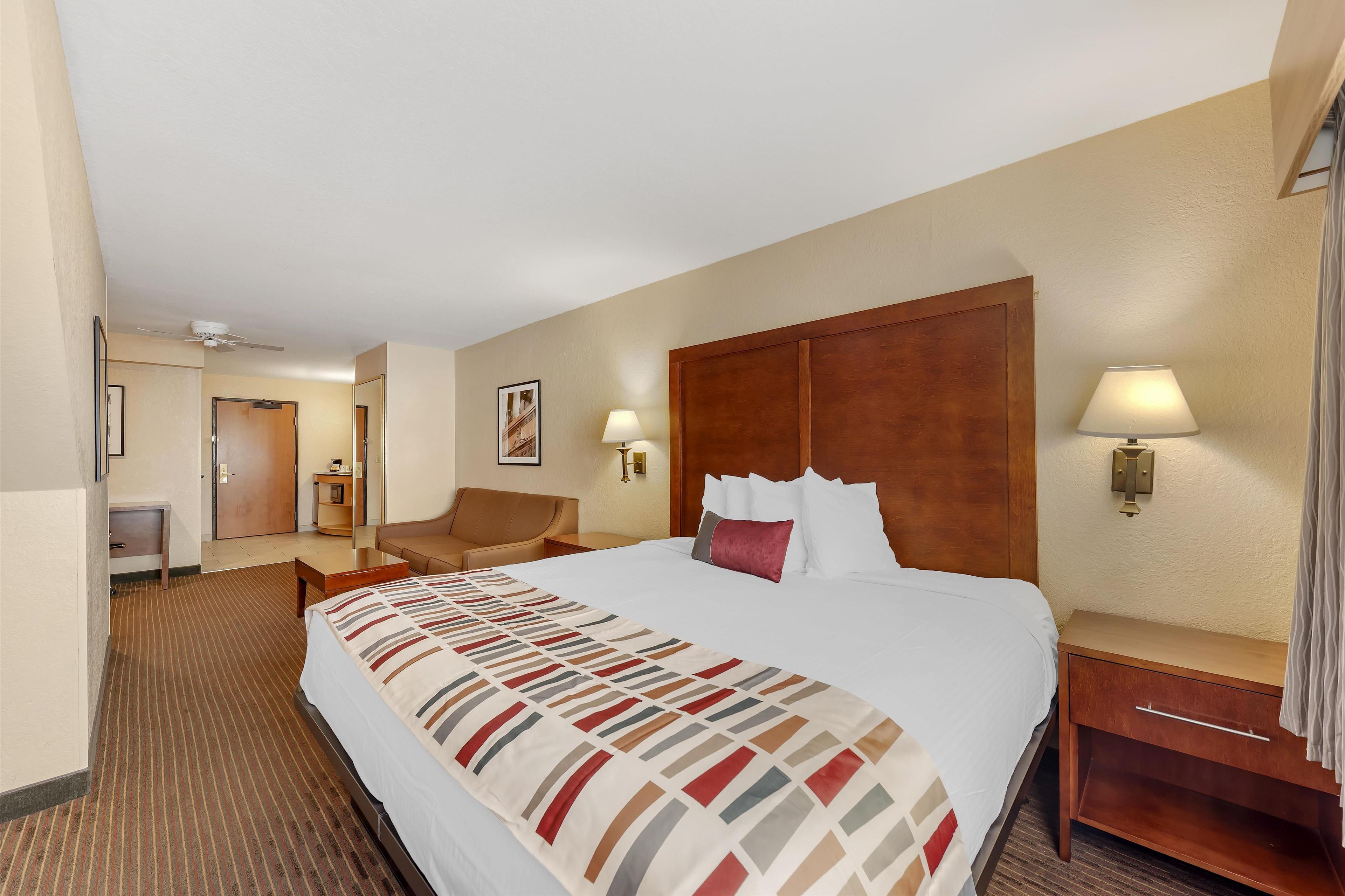 Quarto de hotel com uma cama grande, cabeceira de madeira, mesa de cabeceira com abajur, uma cadeira e uma escrivaninha com luminária.