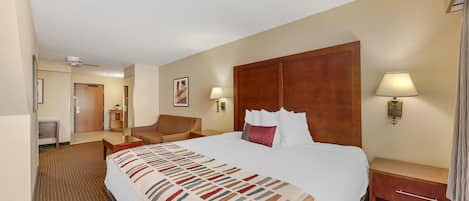 Quarto de hotel com uma cama grande, cabeceira de madeira, mesa de cabeceira com abajur, uma cadeira e uma escrivaninha com luminária.