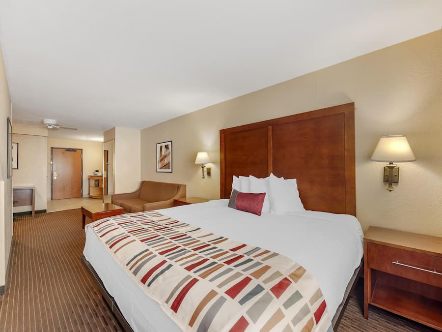 Habitación de hotel con una cama grande, cabecera de madera, mesita de noche con lámpara, una silla y un escritorio con lámpara.