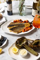 Una mesa de comedor con un plato de pescado a la parrilla, alcachofas y una copa de vino tinto.
