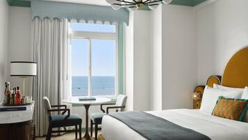 Quarto de hotel com uma cama, uma escrivaninha iluminada por um abajur, uma cadeira, uma mesa e vista para o mar.