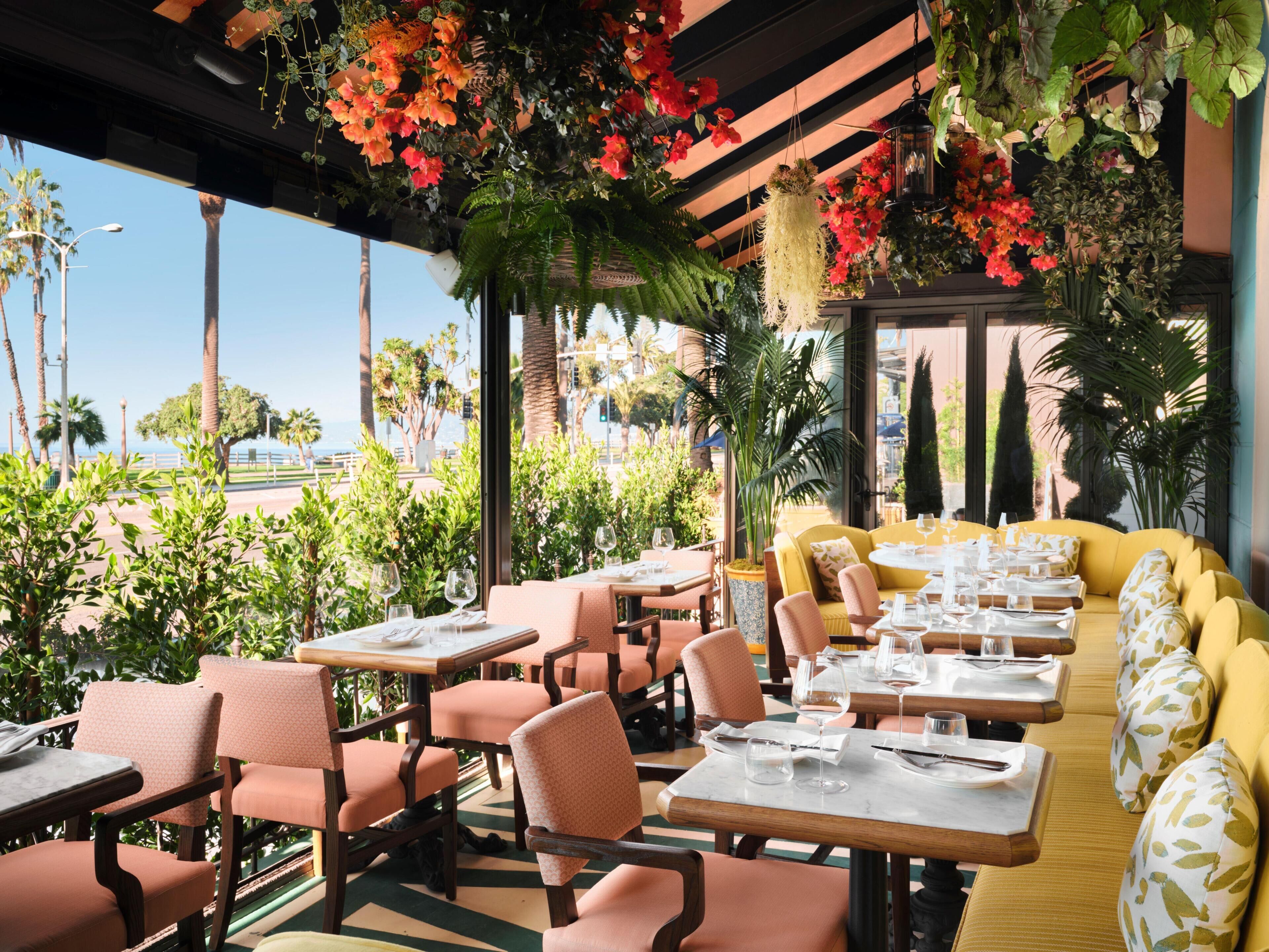 An outdoor dining area with tables and chairs, surrounded by greenery and a view of palm trees.