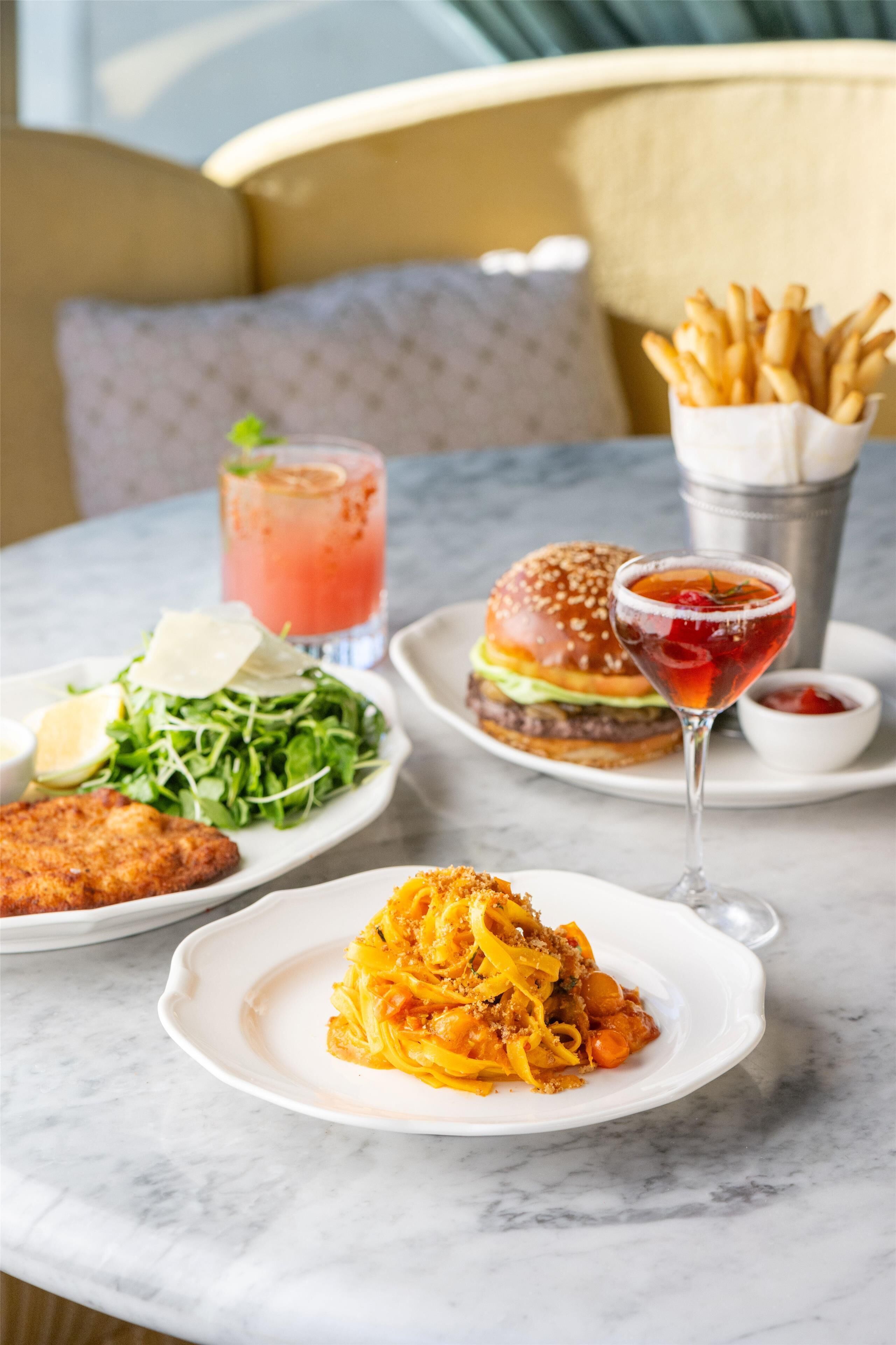 Une table avec différents plats, notamment un hamburger, des frites, une salade et des pâtes, ainsi que deux boissons.