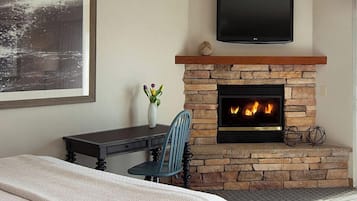 Ein Schlafzimmer mit Kamin aus Stein, einem darüber montierten Fernseher, einem Schreibtisch mit Stuhl und einem Bett mit weißer Bettdecke.