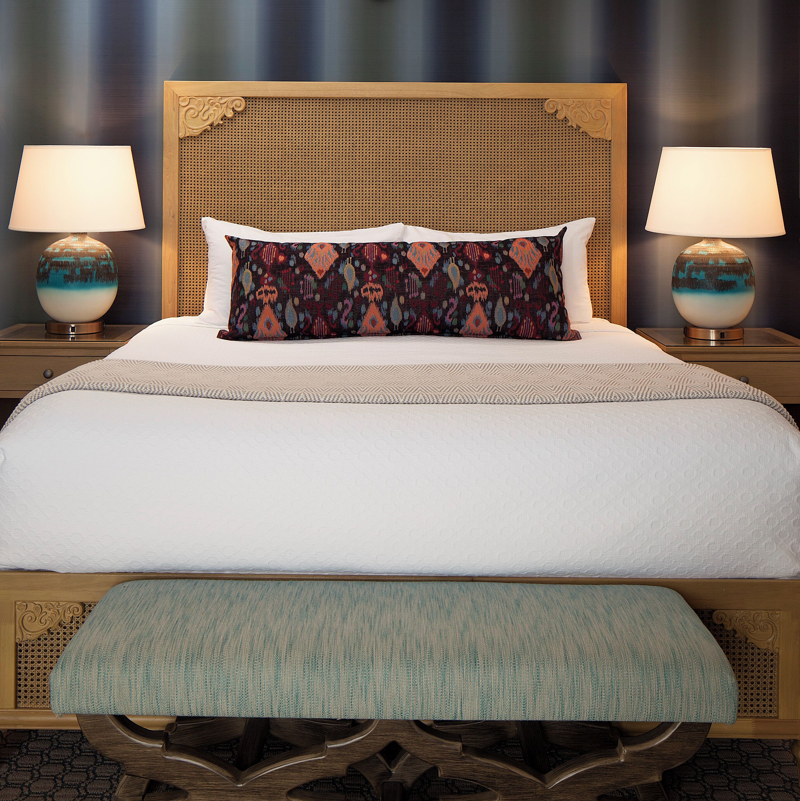 Uma cama bem arrumada com uma almofada estampada, duas luminárias de cabeceira e um banquinho ao pé da cama.