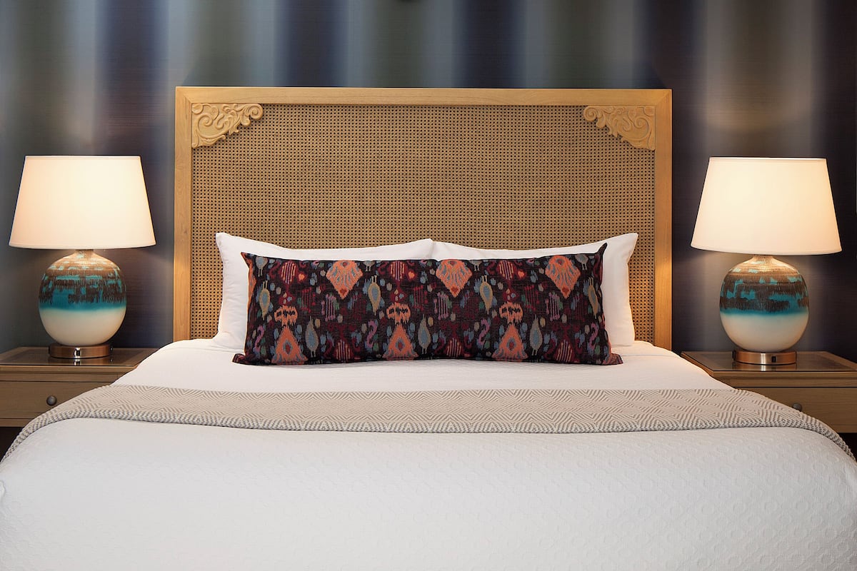 A neatly made bed with a patterned pillow, two bedside lamps, and a bench at the foot of the bed.