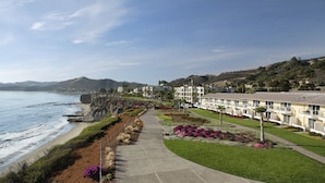 Un hotel costero con un paseo pavimentado a lo largo de la playa, vista al océano y una colina cubierta de vegetación.