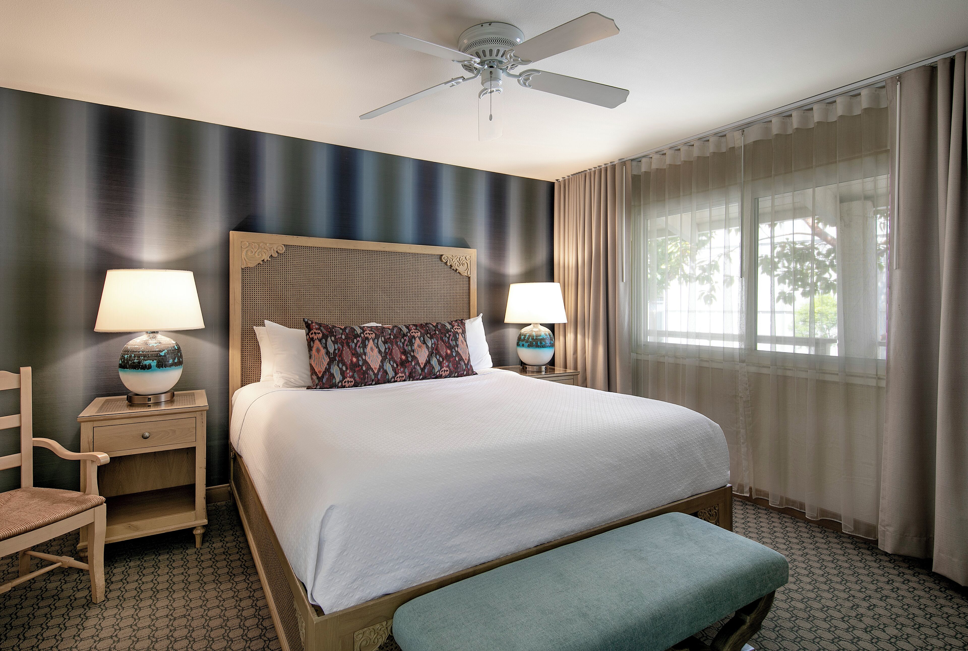 A hotel room with a large bed, two bedside tables with lamps, a ceiling fan, and a window with curtains.