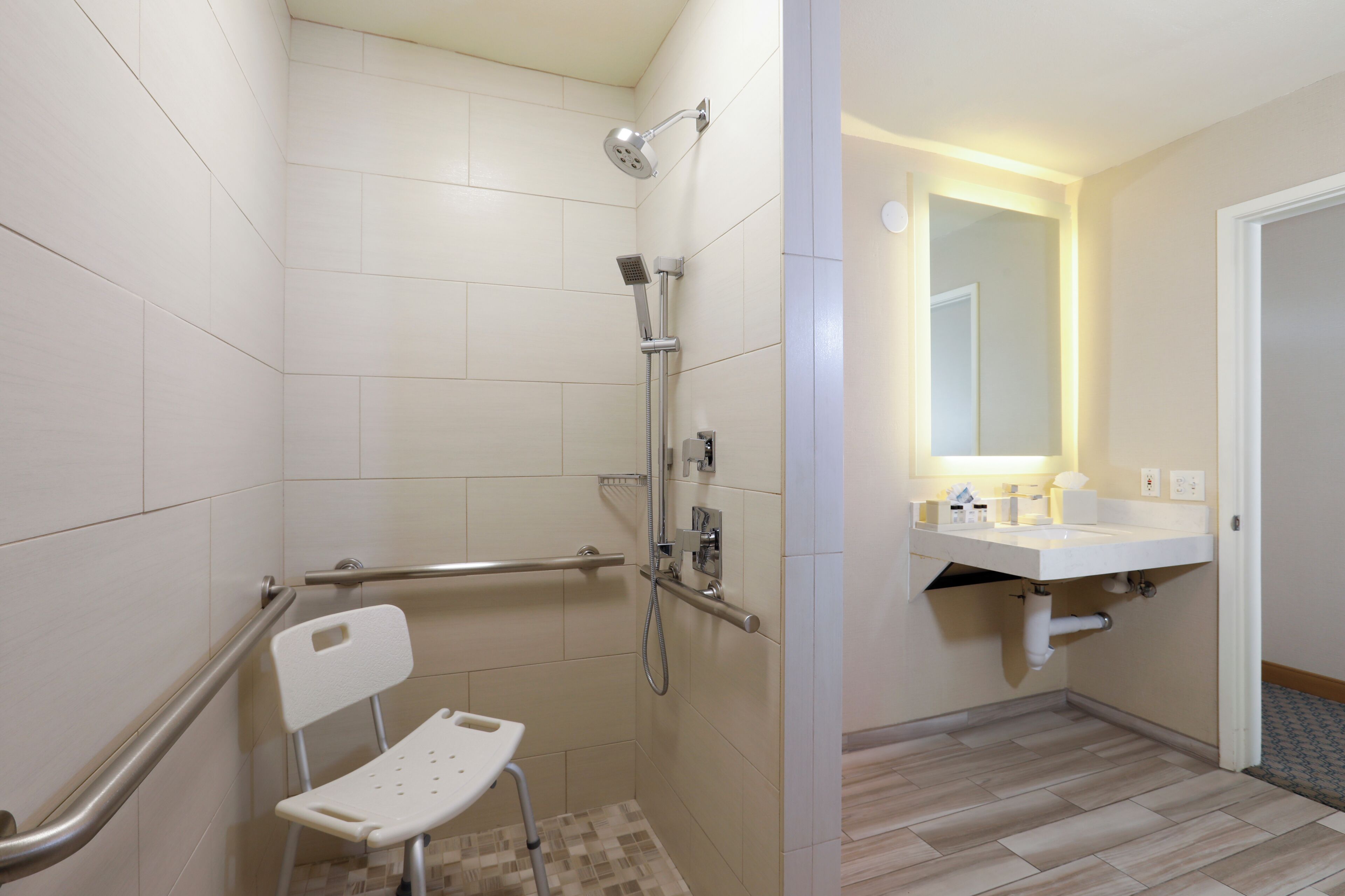 A bathroom with a shower, a handrail, a white chair, and a sink with a mirror.