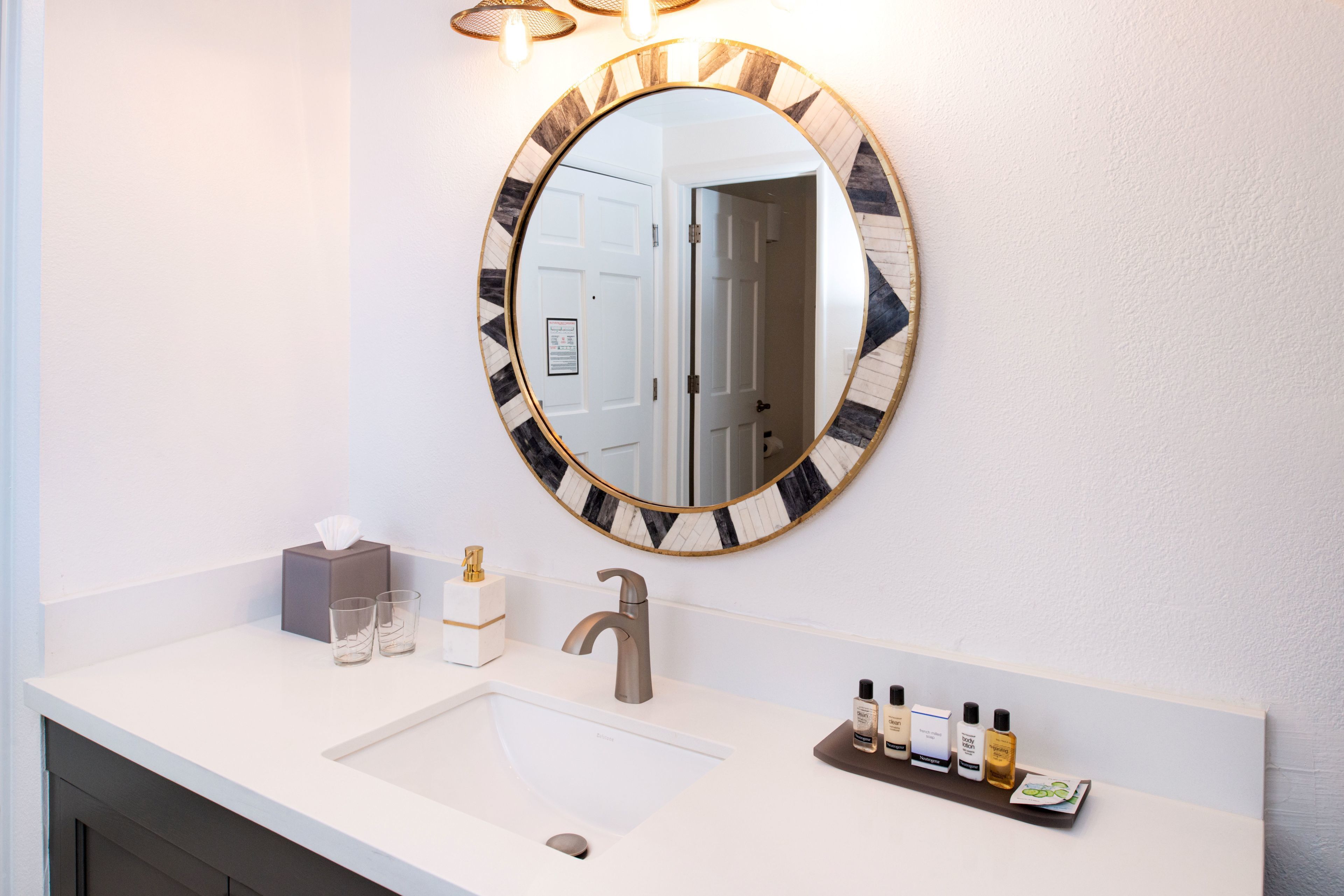 Un baño con un lavabo blanco, un espejo redondo con marco decorativo, un dispensador de toallas y un estante con artículos de aseo.