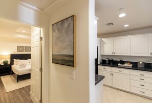A modern kitchen with white cabinets and a black countertop, a bedroom with a bed and nightstands, and a hallway with a painting. - Wine Valley Inn (Solvang)
