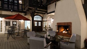 A cozy outdoor seating area with a fireplace, tables, and chairs, surrounded by a brick floor and wooden beams. - Wine Valley Inn (Solvang)