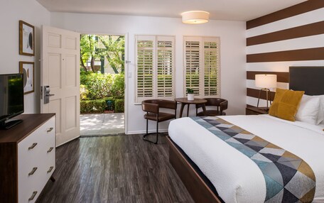 A hotel room with a bed, a desk, a chair, a TV, and a door leading to a balcony with greenery.