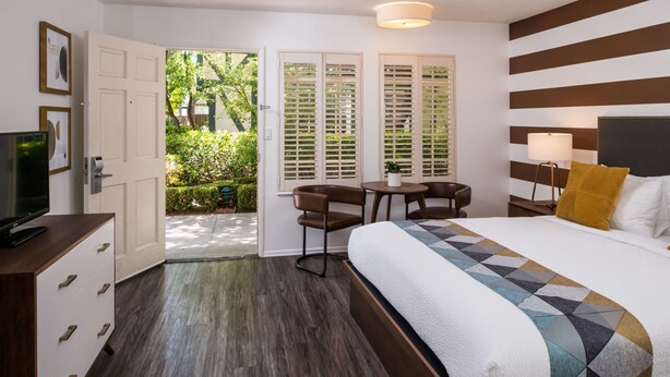 A hotel room with a bed, a desk, a chair, a TV, and a door leading to a balcony with greenery. - Wine Valley Inn (Solvang)