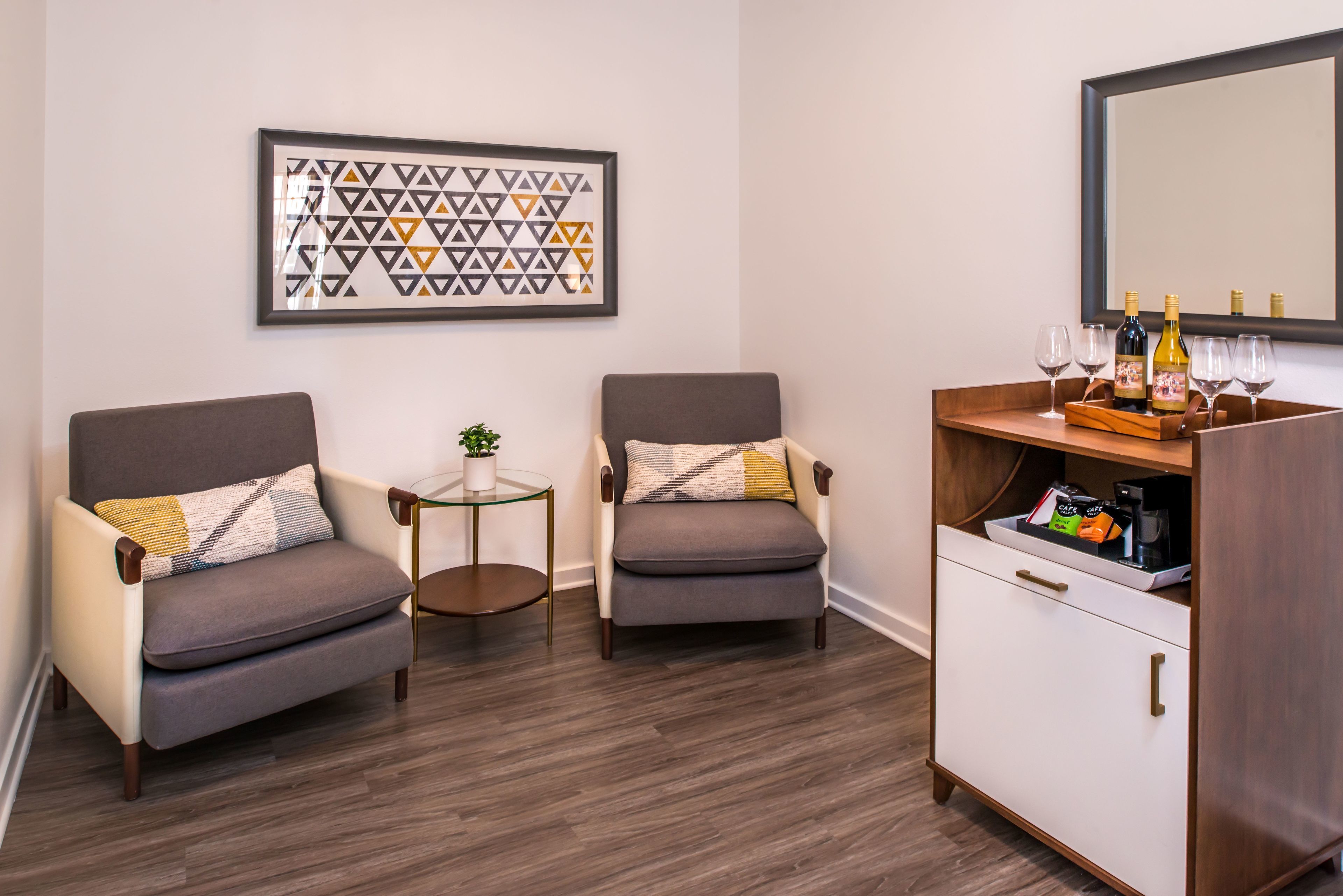 Una habitación moderna con dos sillones, una mesita redonda con una planta, un mueble bar con copas y botellas de vino, y una obra de arte geométrica enmarcada en la pared.