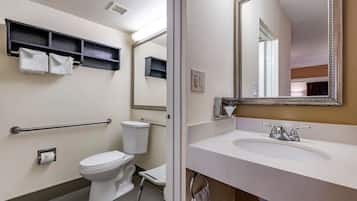 Un baño con inodoro, lavabo y espejo. Toallas cuelgan de un perchero y hay un estante sobre el inodoro.