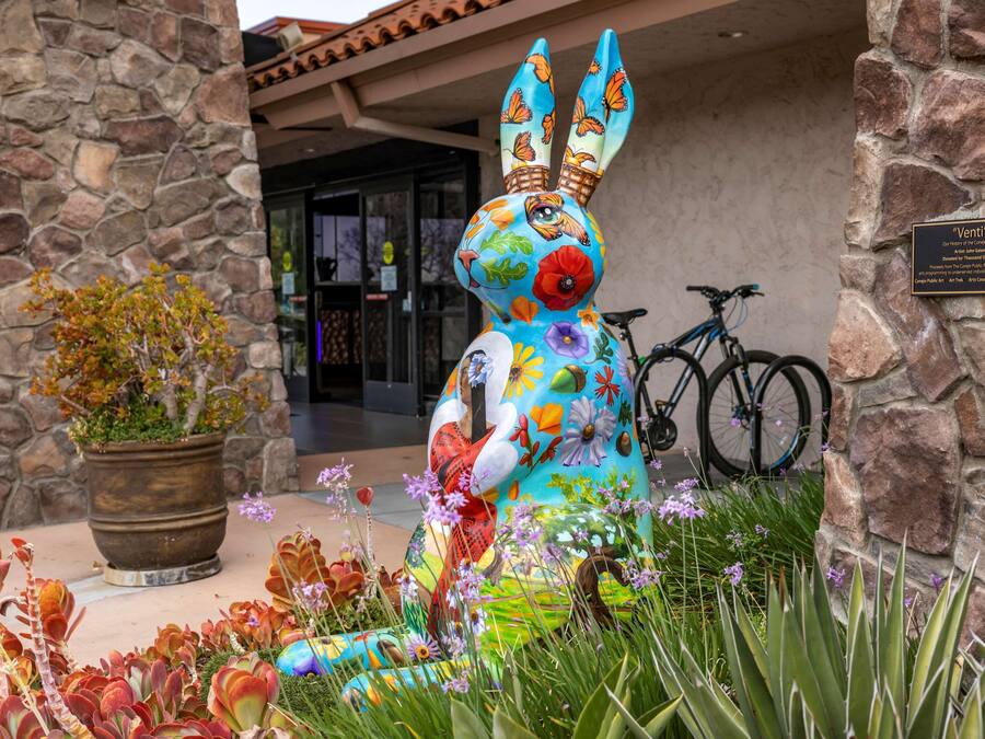Eine farbenfrohe Hasenstatue vor einem Steinhaus, in dessen Nähe ein Fahrrad steht.