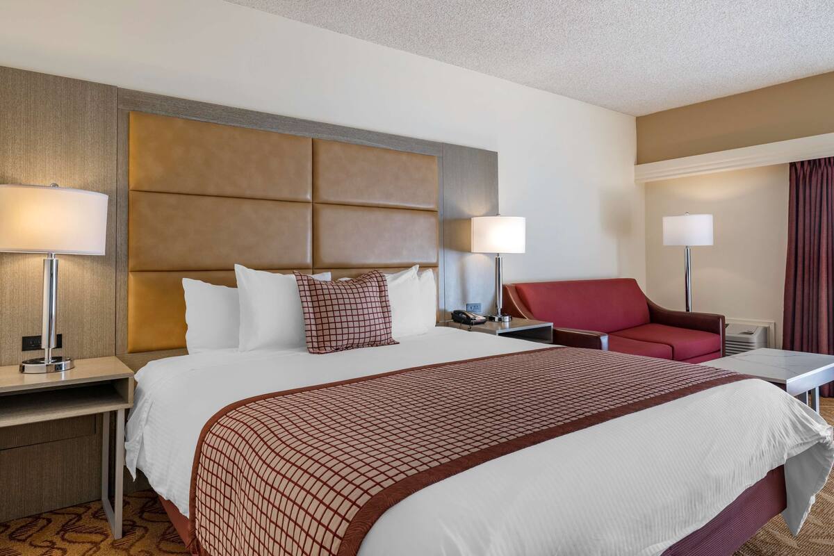 A hotel room with a large bed, two bedside tables, a red armchair, and a desk with a lamp.