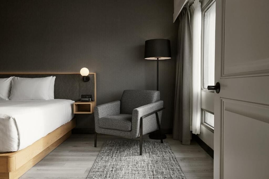 Een hotelkamer met een bed, een stoel, een nachtkastje, een lamp en een raam met gordijnen.