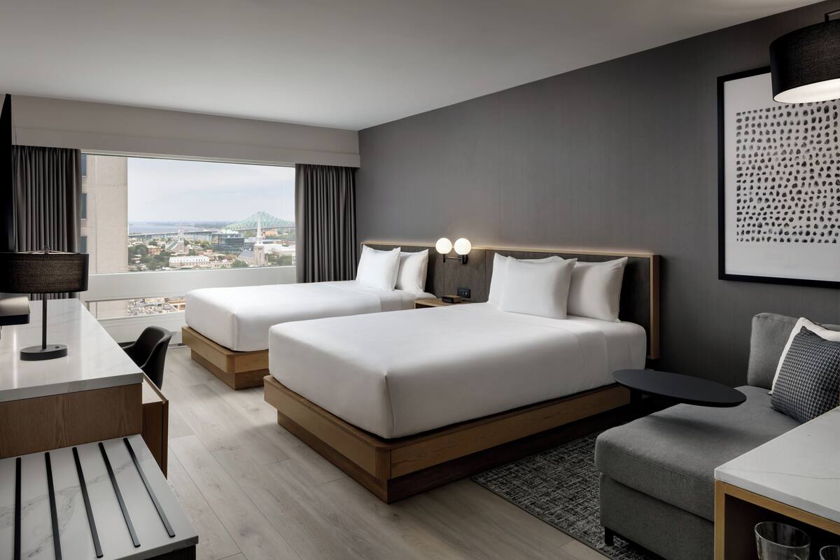 Una habitación de hotel moderna con dos camas, un escritorio, una silla, una mesita y vistas a la ciudad.
