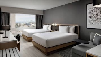 A modern hotel room with two beds, a desk, a chair, a small table, and a view of the cityscape.