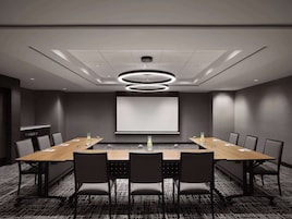 Une salle de conférence équipée d’une longue table, de chaises, d’un écran de projection et d’un luminaire moderne au plafond.