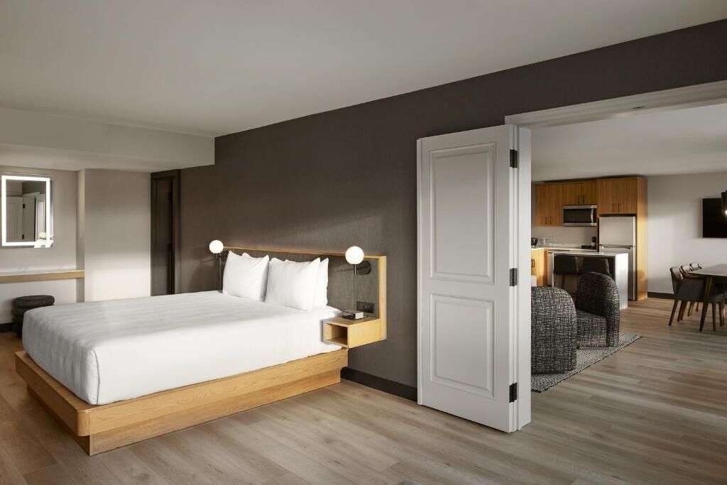 A modern hotel room with a large bed, a desk, and a kitchenette.