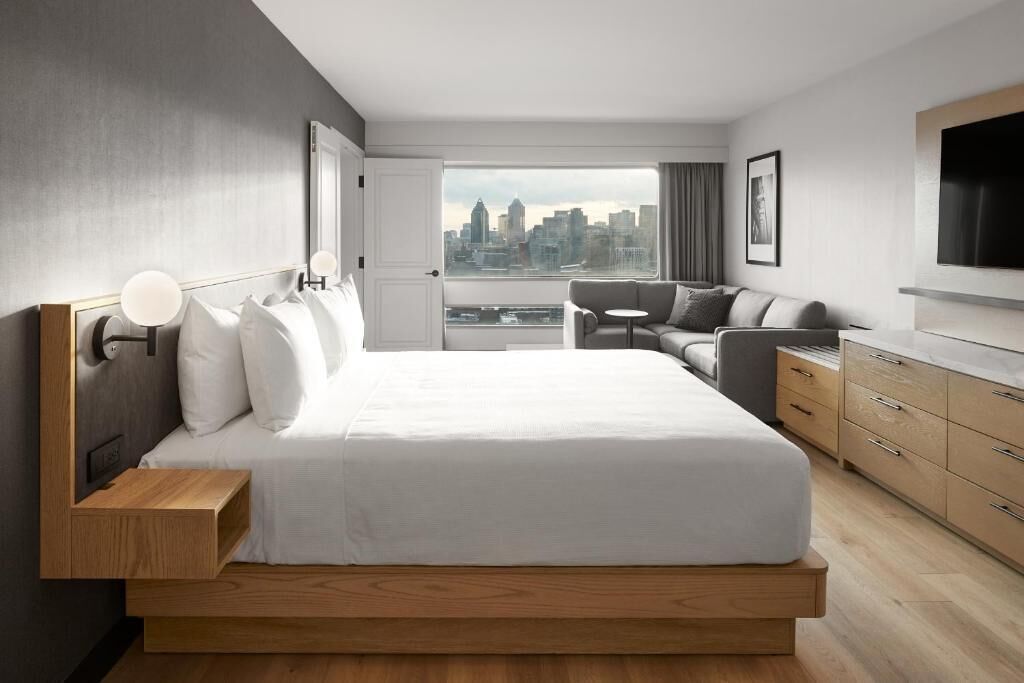 A modern hotel room with a large bed, a sofa, a TV, and a city view.