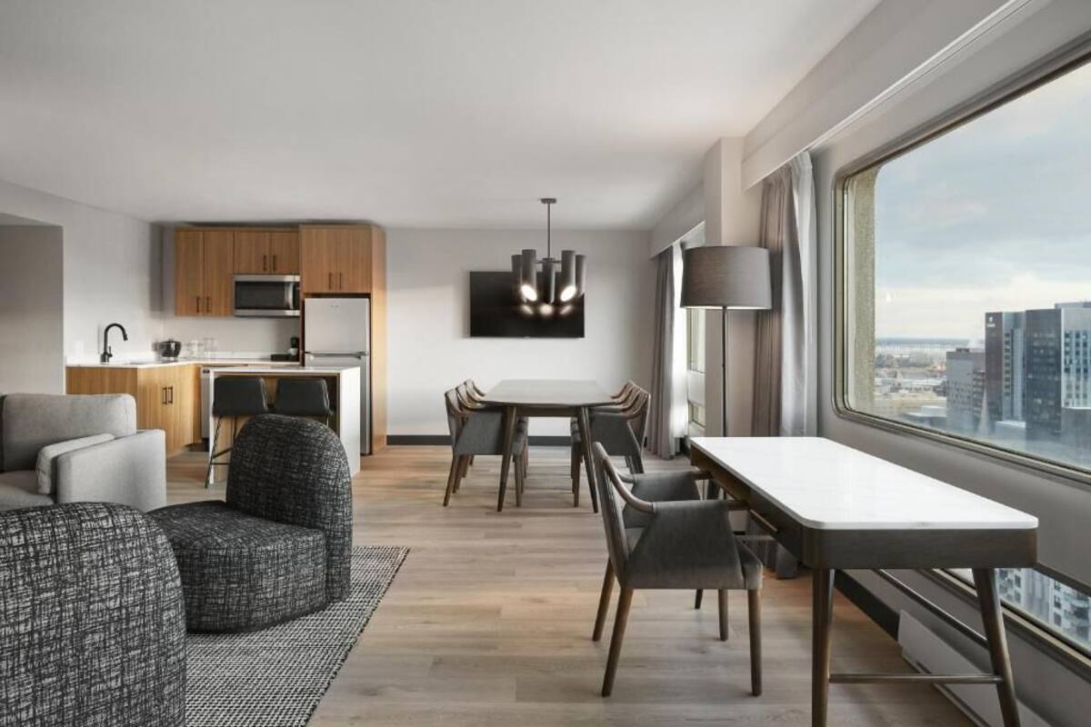 Una habitación de hotel moderna con zona de comedor, kitchenette y un amplio ventanal con vistas a la ciudad.