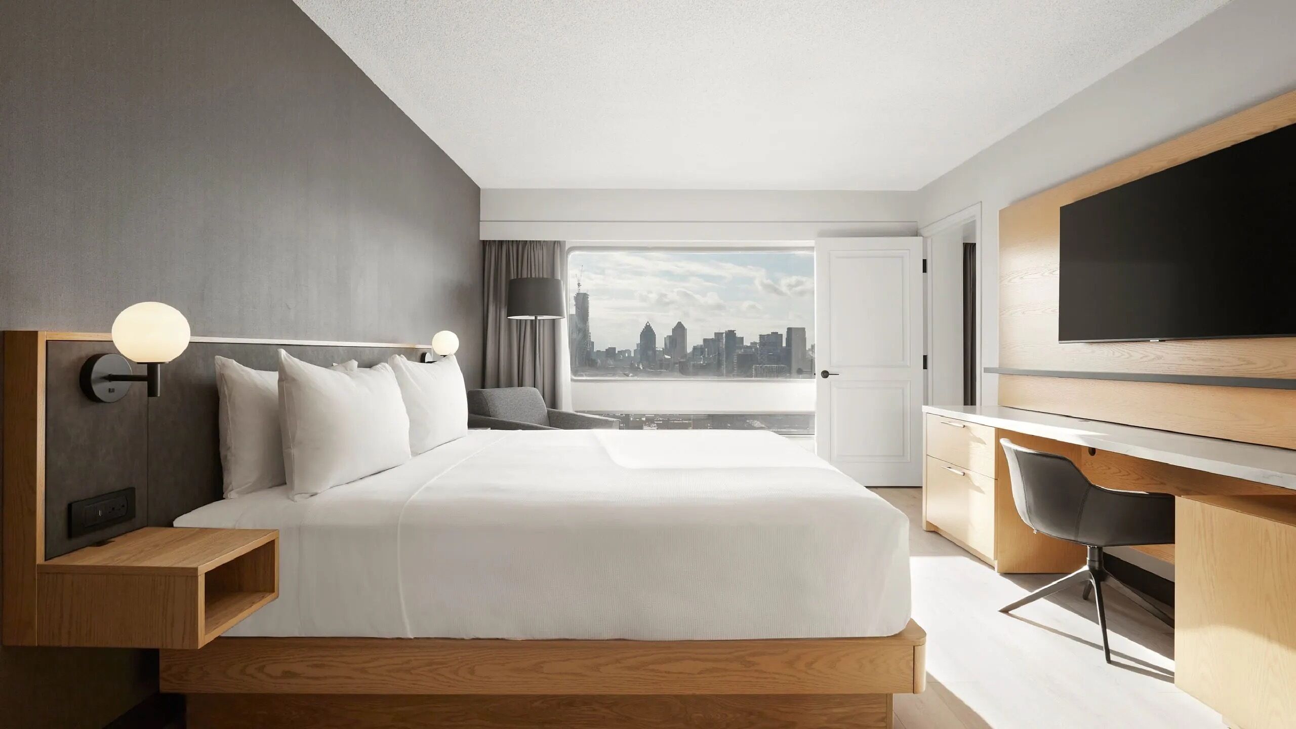 A modern hotel room with a large bed, a desk, a chair, a TV, and a city view.