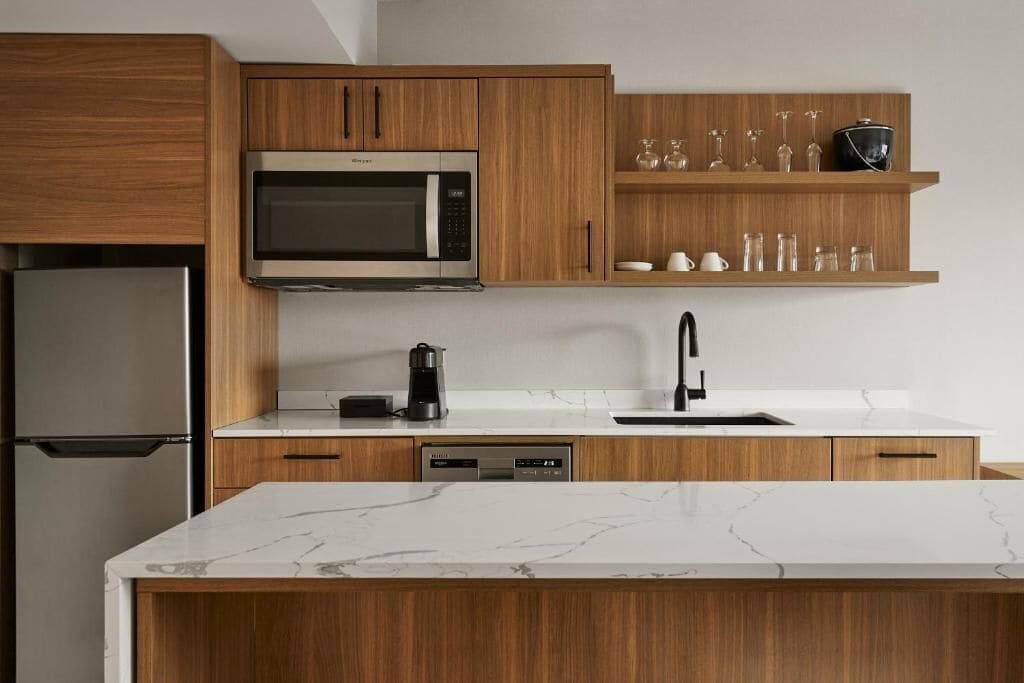 A modern kitchen with wooden cabinets, a built-in microwave, a coffee maker, a dishwasher, and a marble countertop.