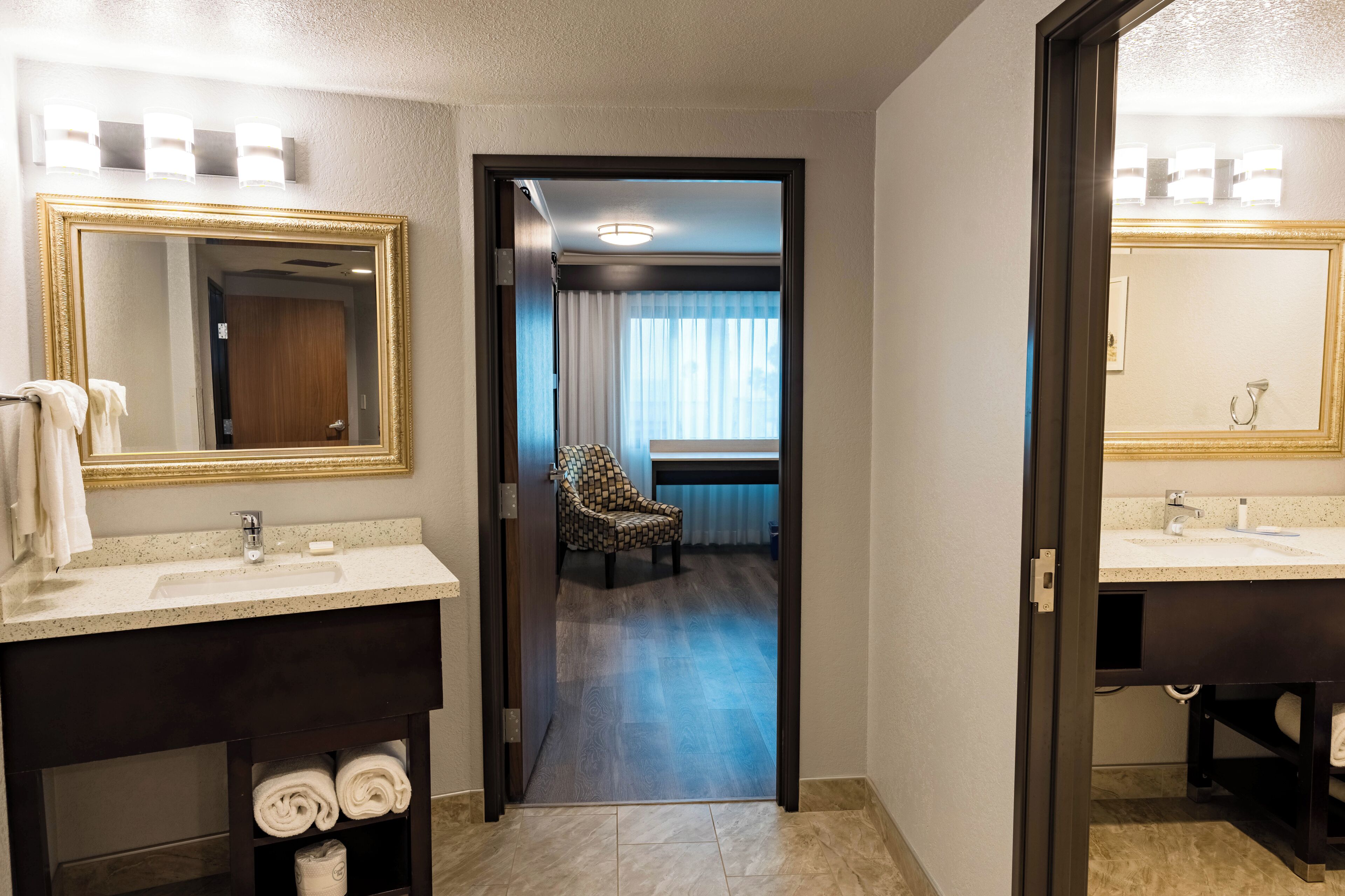 A hotel bathroom with two sinks, a mirror with a gold frame, and a chair in the background.