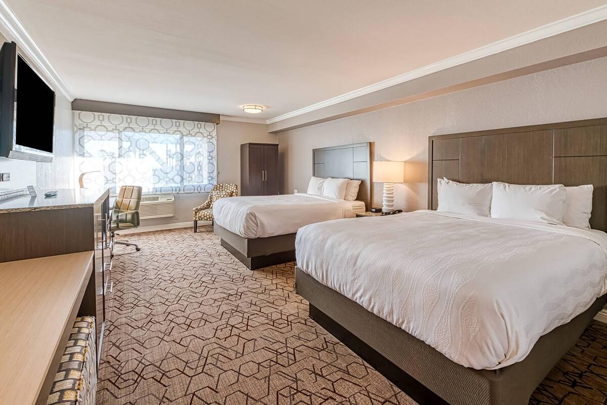 A hotel room with two beds, a desk, a chair, a TV, and a window with patterned curtains.