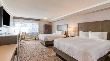A hotel room with two beds, a desk, a chair, a TV, and a window with patterned curtains.