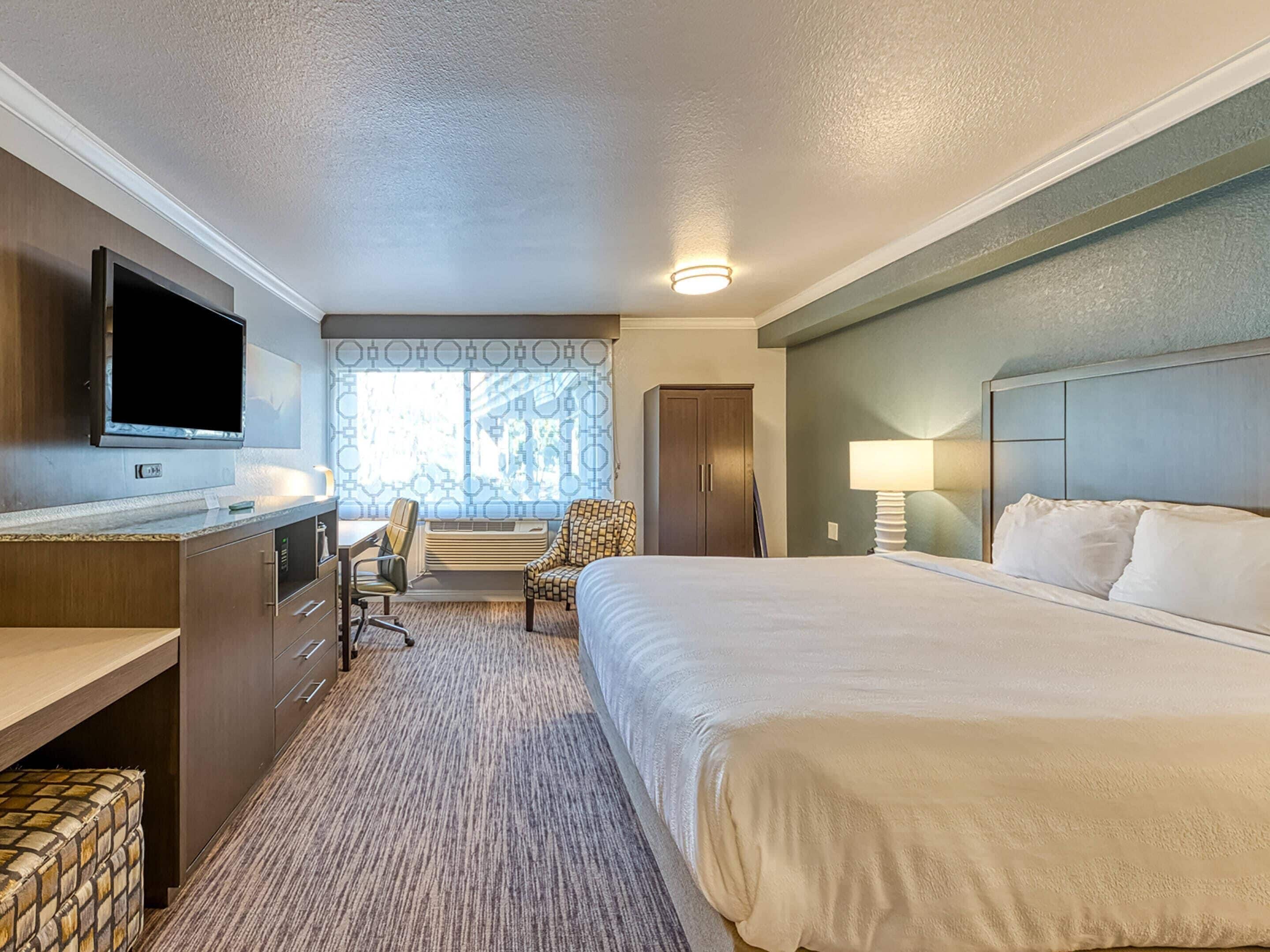 Habitación de hotel con cama, escritorio, silla, televisor y ventana con cortinas.