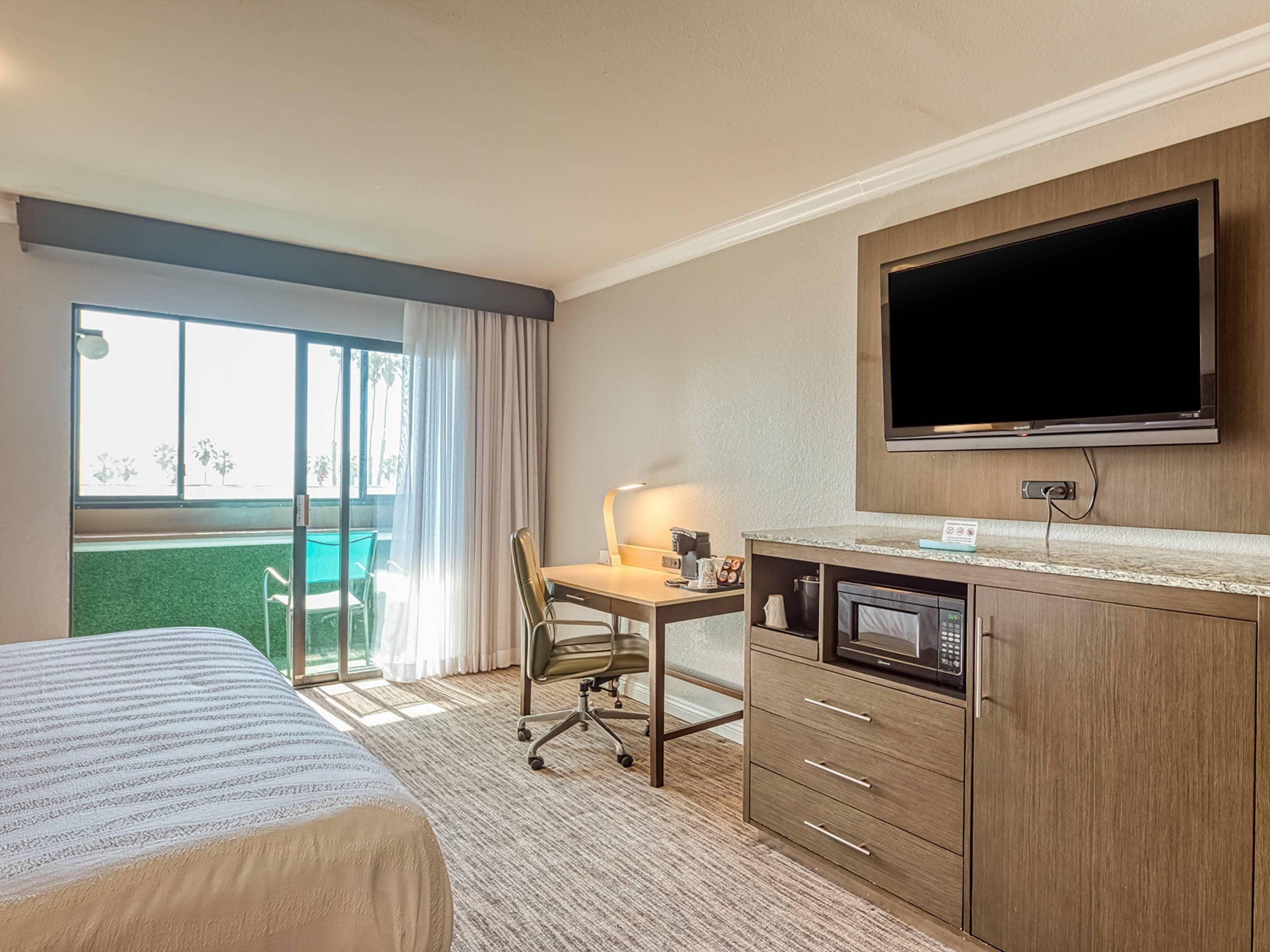A hotel room with a bed, desk, chair, television, and a view of a green outdoor area.